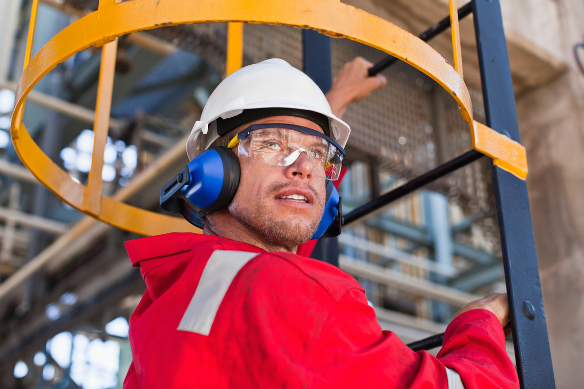 a person wearing personal protective equipment smiles while climbing a ladder in an industrial setting