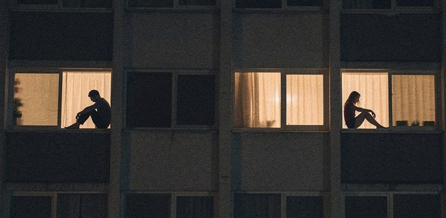 If Youre Suffering And Questioning God, Youre Not Alone 1 2026 01 26 Two People Sitting Alone In Separate Lit Apartment Windows At Night Building Facade Isolation Loneliness Urban Solitude e1770393145468
