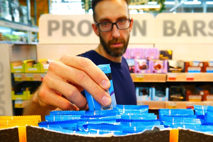 639138248006011925man picking up protein bar in supermarket